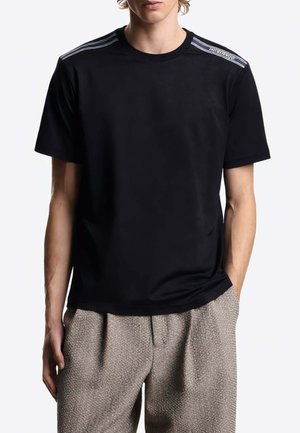 Uomo che indossa una t-shirt nera con scollo rotondo e dettaglio a righe sulle spalle e pantaloni beige testurizzati, con le mani nelle tasche.