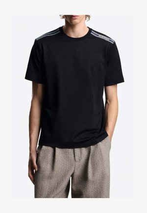 Uomo che indossa una t-shirt nera con scollo rotondo e dettaglio a righe sulle spalle e pantaloni beige testurizzati, con le mani nelle tasche.