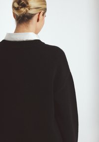 Black knit sweater with a high neckline and dropped shoulders, featuring a white collared shirt underneath. Soft texture, minimalistic design.