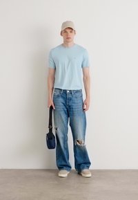 Hellblaues T-Shirt mit kurzen Ärmeln, zerissenes hellblaues Jeans, beiger Hut, cremefarbene Slipper und eine kleine marineblaue Umhängetasche.