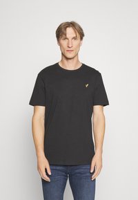 Schwarzes T-Shirt mit kurzen Ärmeln aus weichem Stoff, mit Rundhalsausschnitt und einem kleinen goldenen Logo auf der linken Brust.
