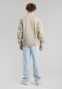 Beige sweatshirt with high collar, loose fit, and ribbed cuffs. Worn with light blue jeans and white sneakers. Plain and smooth texture.
