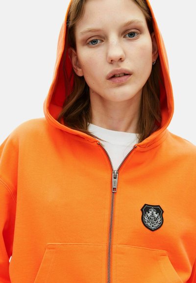 Felgekleurde oranje zip-up hoodie van zacht katoen, met een voorzak en een zwart embleem op de linker borst. Soepele stoftextuur.