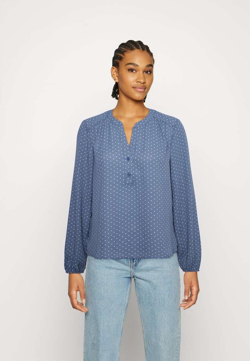 Blusa azul de manga longa com gola henley abotoada, com bolinhas brancas e ombros franzidos, combinada com jeans de lavagem clara.