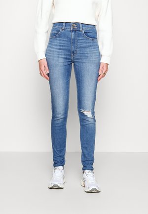 High-waisted blue skinny jeans in denim, featuring a distressed knee, five-pocket design, and subtle stretch fabric. Paired with white sneakers.