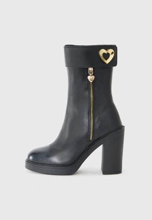 Bottine en cuir noir avec un haut talon bloc, fermeture éclair sur le côté et un accent en métal doré en forme de cœur sur le revers. Texture lisse.