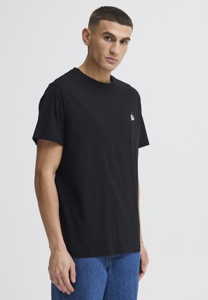 Ung mand iført en almindelig sort t-shirt med et lille broderet design på brystet og blå jeans, stående mod en hvid baggrund.