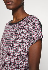 Mönstrad topp med ett rosa hjärtmotiv på en svart bakgrund, med svart halsringning och korta ärmar. Slät tygstruktur.