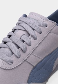 Κοντινό πλάνο ανοιχτόγκρι αθλητικού παπουτσιού με υφασμένες κορδόνια και ένα μπλε-γκρι κεντημένο λογότυπο στο πλάι.