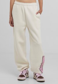 Crème kleurige sweatpants met een elastische tailleband, zijzakken en een roze geborduurd detail op het linkerpand. Zacht, ontspannen pasvorm.