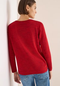 Maglione rosso lavorato a maglia con maniche lunghe, scollo a giro e finitura testurizzata. Abbinato a jeans in denim blu, visto da dietro contro un muro.