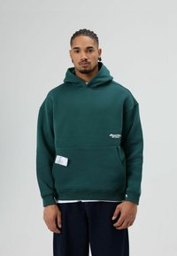 Donkergroene hoodie met een voorzak en witte logodetails. Heeft een losse pasvorm, geribbelde manchetten en een zachte stoftextuur.