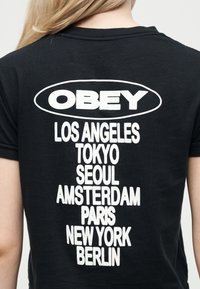 Άτομο που φοράει μαύρο μπλουζάκι με το λογότυπο «OBEY» πάνω από μια λίστα πόλεων: Λος Άντζελες, Τόκιο, Σεούλ, Άμστερνταμ, Παρίσι, Νέα Υόρκη, Βερολίνο.
