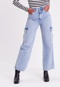 Lichtblauwe denim cargobroek met brede pijpen, twee zijzakken, hoge taille en een vervaagde afwerking. Zwarte enkellaarsjes erbij gedragen.