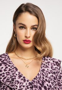 Violetti leopardi kuvioitu mekko, jossa on röyhelökaulus, kultaketju kaulaketjut ja renkaakorviksia. Sileä tukka muotoiltuna löysille laineille.