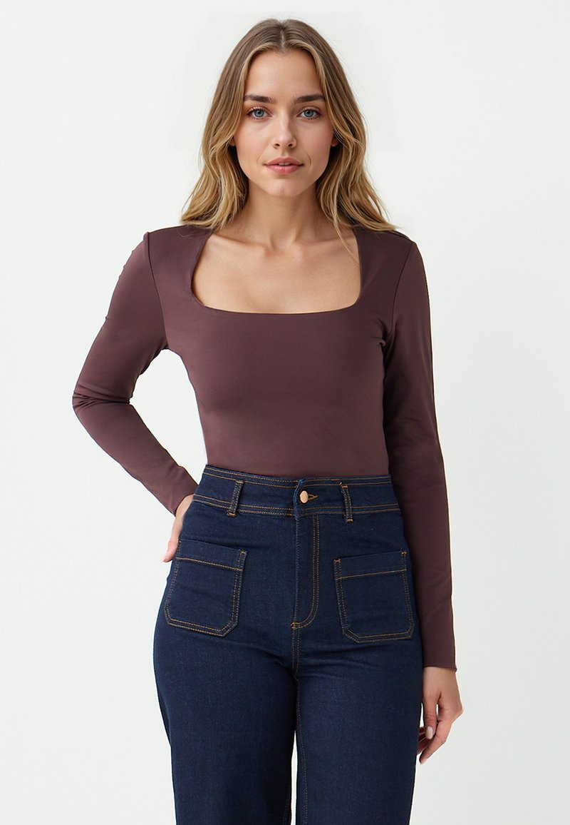 Braunes Langarmoberteil mit quadratischem Ausschnitt, kombiniert mit hochgeschnittenen dunkelblauen Jeans, die sichtbare Nähte und Taschen vorne aufweisen.