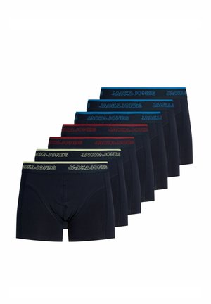 Mladý muž s krátkymi blond vlasmi, oblečený v čiernych boxerkách Jack & Jones, stojaci pred jednoduchým svetlým pozadím.