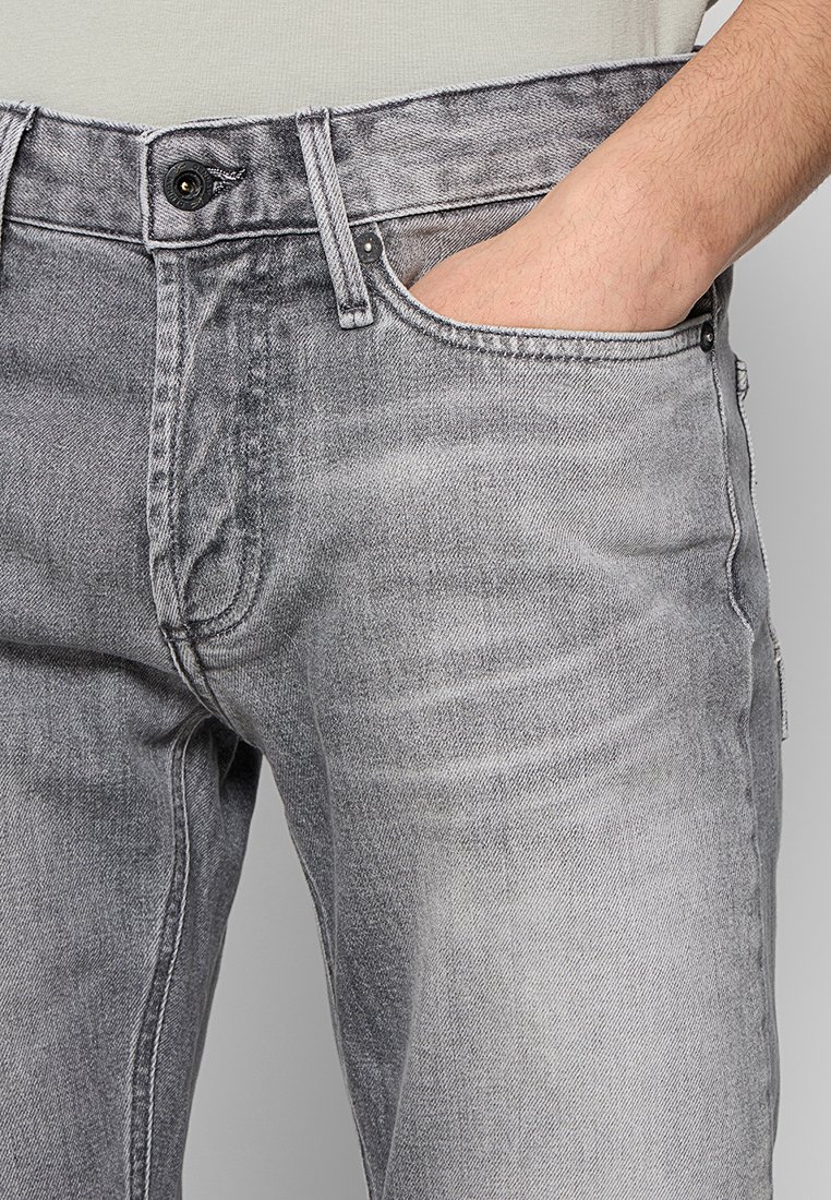 Hand in de zak van grijze denim jeans met zichtbare knoop, ritssluiting, riemlussen en stikseldetails.