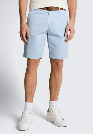 Man die lichtblauwe knielange shorts, een bruine riem, witte sokken en witte sneakers draagt en staat tegen een effen achtergrond.