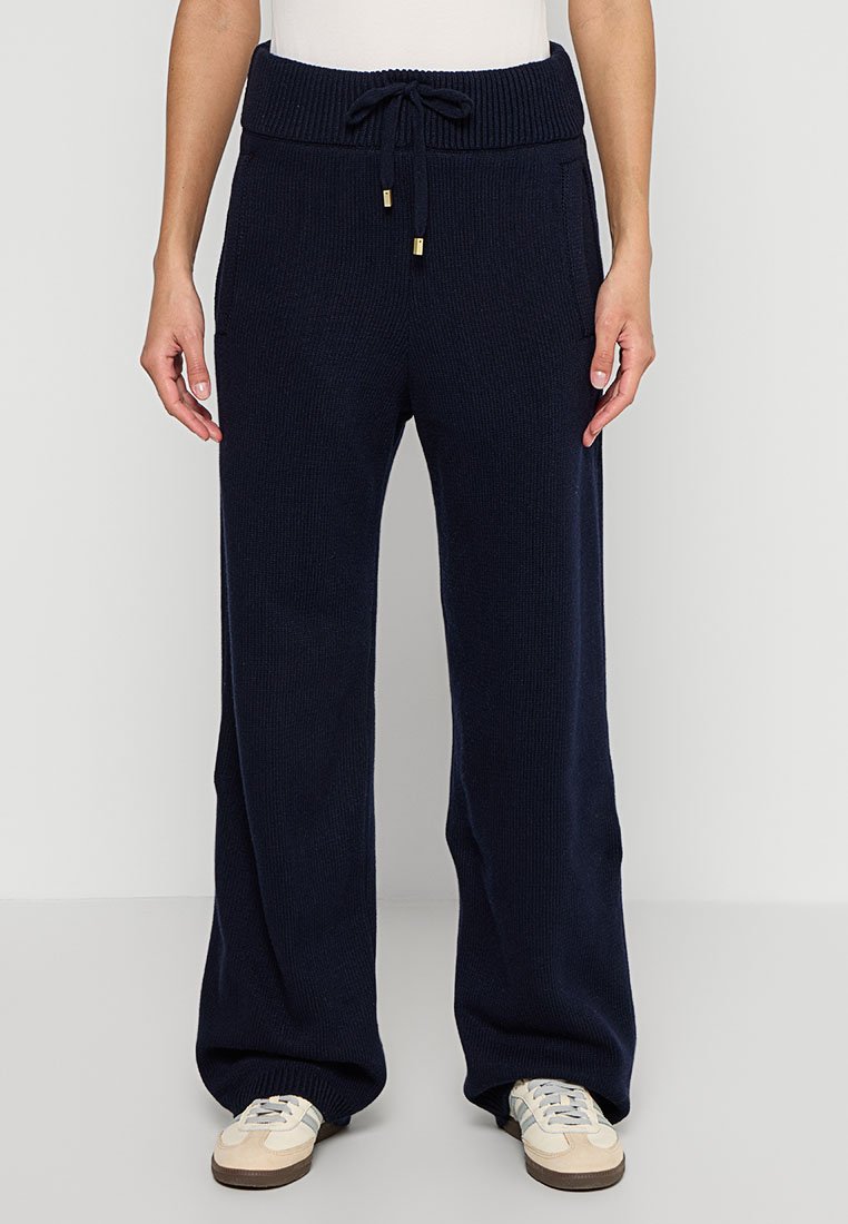 Pantalones de punto acanalado en navy con cintura ajustable, con detalles de herrajes dorados y pierna ancha. Combinados con zapatillas beiges.