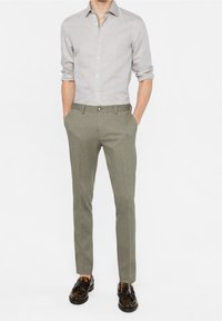 Ljusgrå skjorta med knappar, slim-fit olivgrön ullbyxor och mörkbruna läderskor med glänsande yta, händerna i fickorna.