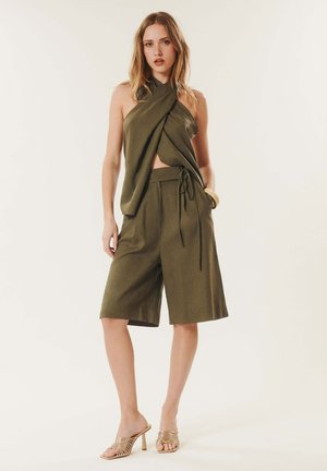Femme portant un top dos nu vert olive et un short large assorti longueur genou avec des sandales à talons hauts dorées sur un fond uni.