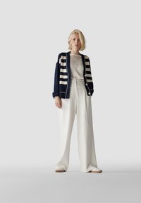 Marineblau- und cremegestreifter Cardigan über einer hellen Bluse, kombiniert mit weißen, weit geschnittenen Hosen und hellen Schuhen, vor einem grauen Hintergrund.