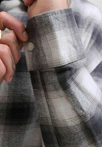 Puño de camisa de franela a cuadros en gris y blanco, con un botón de plástico y una tela texturizada. Costuras visibles y un acabado suave.
