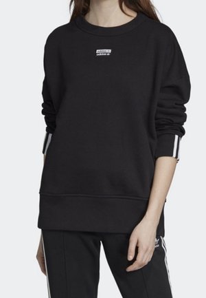 Černá mikina Adidas s dlouhým rukávem, malým logem na hrudi a pruhovanými detaily na manžetách, nošená s odpovídajícími černými kalhotami Adidas.