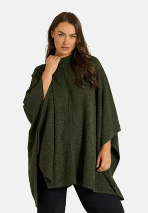 Femme aux longs cheveux bruns portant un poncho en tricot vert foncé et un pantalon noir, debout devant un fond clair uni.
