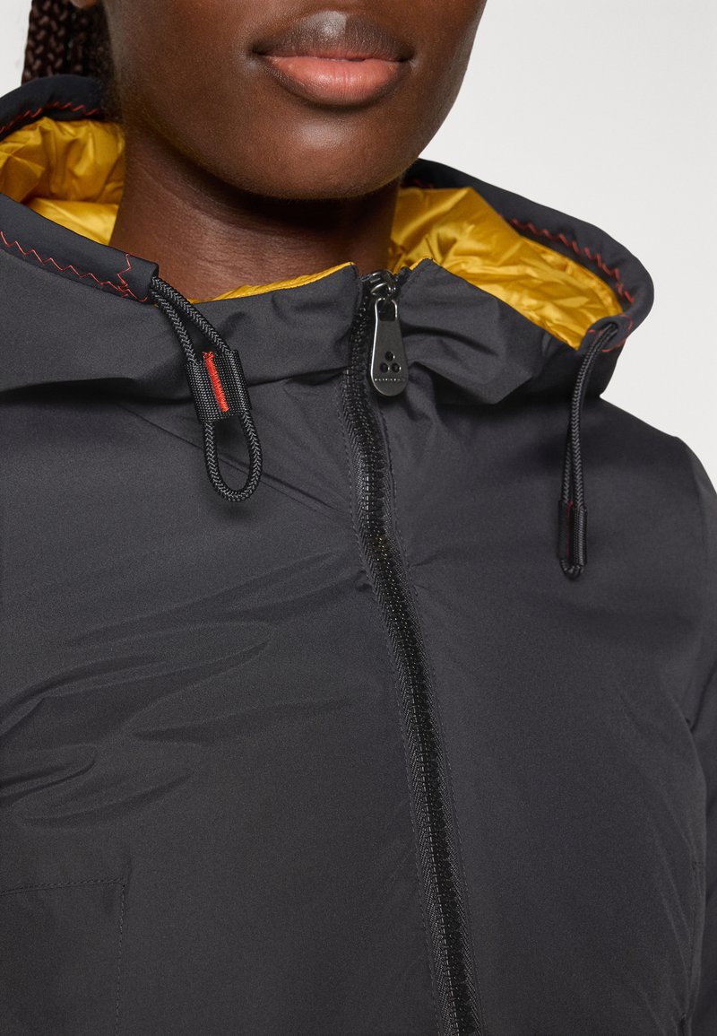 Nahaufnahme einer Person, die eine schwarze Jacke mit gelber Kapuzenfutter trägt, die zugezippt ist und schwarze Kordeln mit roter Naht hat.