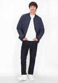 Marine bomberjack met witte ankerprint, ribgebreide manchetten en tailleband. Gedragen over een wit T-shirt, gecombineerd met donkere jeans en witte sneakers.