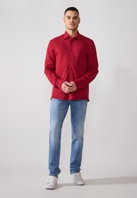 Camisa roja de manga larga con cuello puntiagudo y botones en el frente, combinada con jeans de mezclilla azul claro y zapatillas blancas.