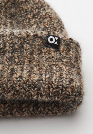 Berretto lavorato a maglia in tonalità di marrone con un aspetto texturizzato e peloso. Presenta un'etichetta nera con un logo bianco sul lato.