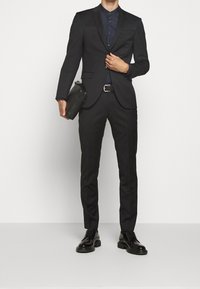 Zwart op maat gemaakt pak met slim-fit broek, eenkoloms colbert en donkerblauw overhemd. Accessoires omvatten een zwarte leren tas en zwarte schoenen.