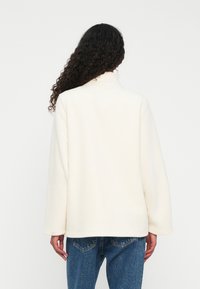 Donna con lunghi capelli ricci che indossa una giacca in pile di colore crema dal taglio ampio e jeans blu, vista di spalle su uno sfondo neutro.