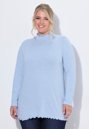 Femme souriante aux cheveux blonds porte un haut à manches longues bleu clair avec des bords volantés et un jean bleu foncé sur un fond blanc uni.