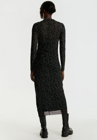 Schwarzes langärmliges Kleid mit weißen und blauen Punkten, taillierte Silhouette, strukturierter Stoff und ein Schlitz hinten am Saum. Dazu werden schwarze Stiefel getragen.