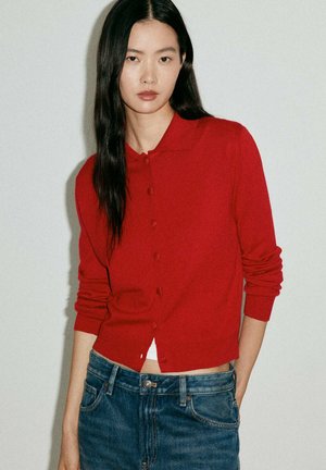 Mujer con cabello largo y negro que lleva un cárdigan rojo con botones y vaqueros azules sobre un fondo sencillo y claro.