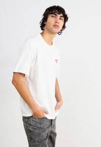 Hvid bomulds t-shirt med korte ærmer, prydet med et lille rødt logo på brystet. Båret med grå mønstrede denimjeans.