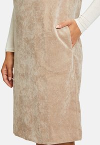 Närbild av en person i en beige texturerad klänning med en hand i sidofickan och långa ärmar i krämf ärg synliga.