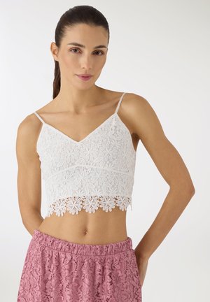 Donna con capelli scuri, indossa un top corto in pizzo bianco con motivi floreali e pantaloncini a vita alta in pizzo rosa, in piedi davanti a uno sfondo bianco uniforme.