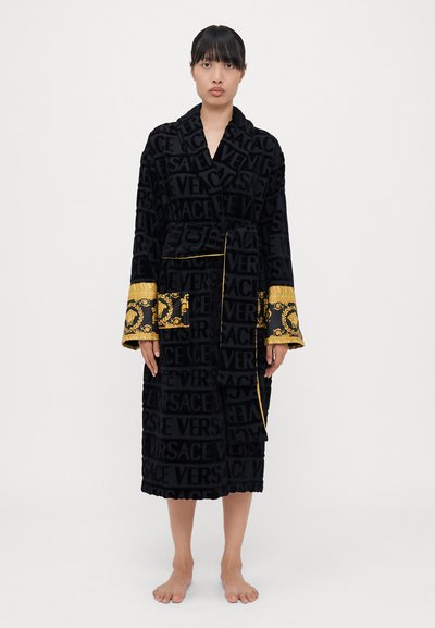 Kvinde iført en sort Versace badekåbe med manchetter og bælte i guld-barok mønster, stående barfodet mod en ensfarvet hvid baggrund.