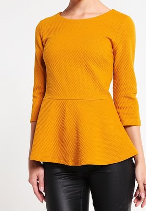 Kvinde iført en tekstureret, sennepsgul peplum-top med trekvartlange ærmer og sorte, tætsiddende bukser.