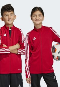 Rote sportliche Zip-Jacken mit weißen Streifen an den Ärmeln, aus glattem Stoff. Ein Kind hält einen gemusterten Fußball, beide in schwarzen Hosen.