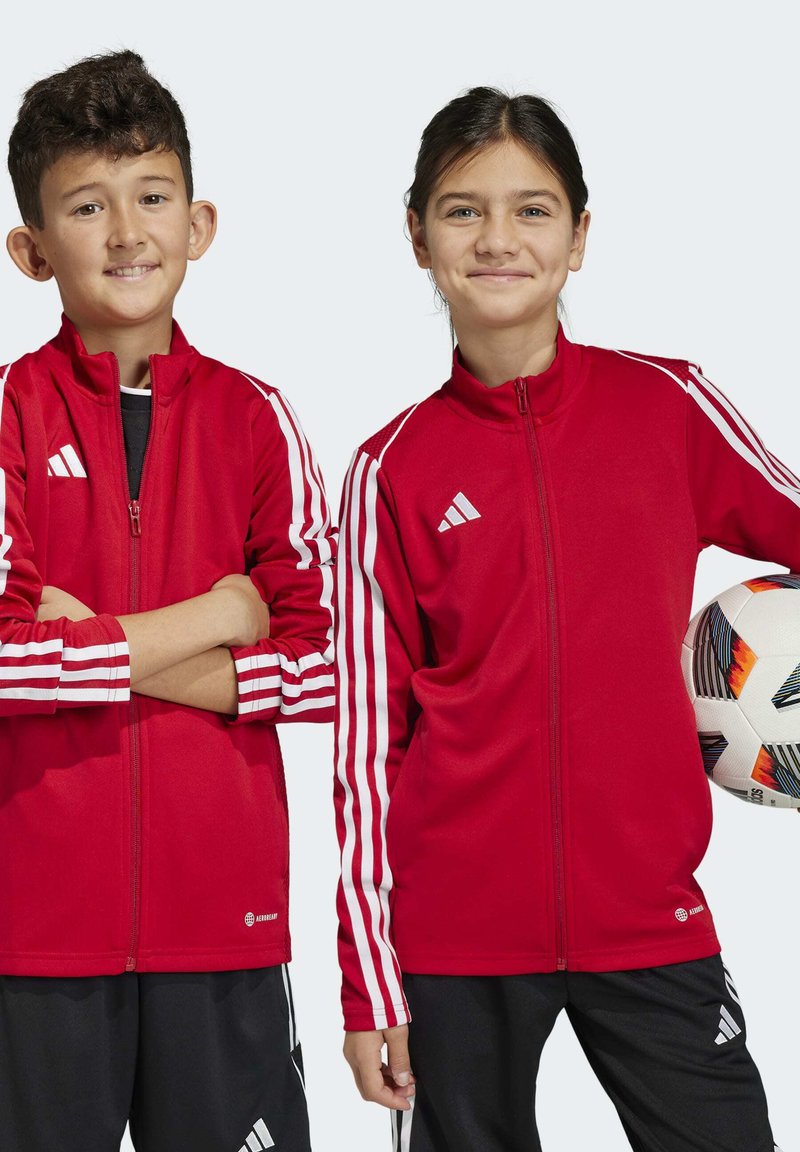 Rote sportliche Zip-Jacken mit weißen Streifen an den Ärmeln, aus glattem Stoff. Ein Kind hält einen gemusterten Fußball, beide in schwarzen Hosen.