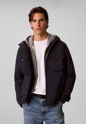 Ung mand med brunt hår iført en sort jakke over en grå lynlåshættejakke og hvid t-shirt, kombineret med blå jeans og et sort bælte.