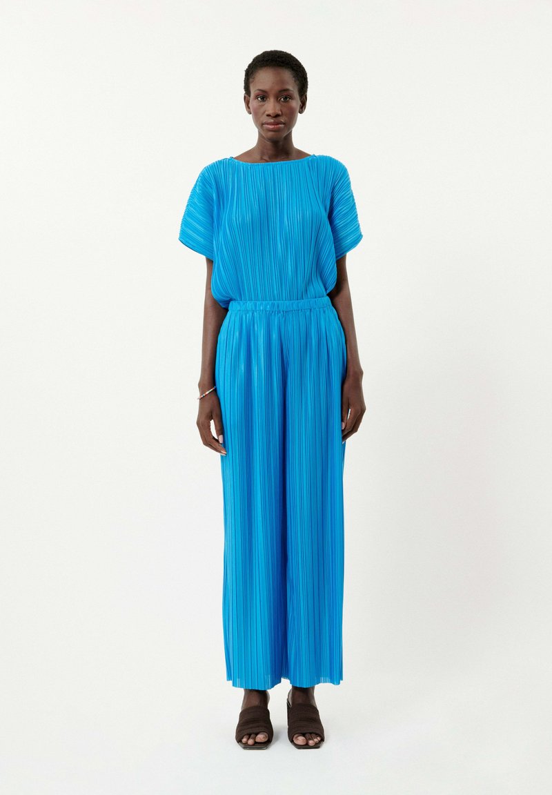 Combinaison bleue plissée avec manches courtes, taille élastique et jambes larges. Texture lisse et couleur vive, agrémentée de sandales brunes.