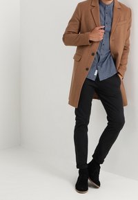 Cappotto di lana marrone, design aderente, monopetto con rever a incavo. Indossato sopra una camicia blu con motivo e abbinato a pantaloni e scarpe nere.