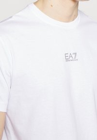 Nahaufnahme einer Person, die ein weißes T-Shirt mit einem kleinen EA7 Emporio Armani-Logo auf der linken Brust trägt.